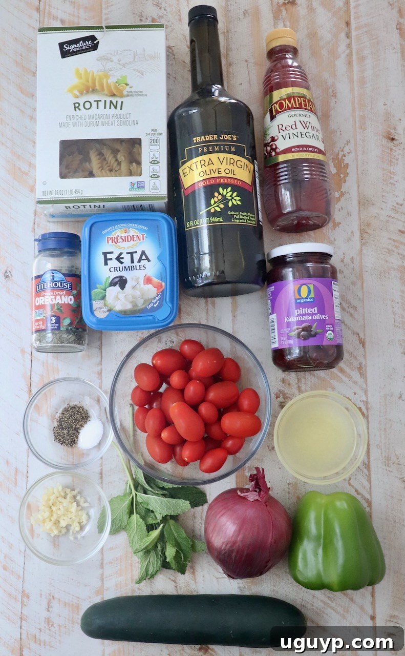 Fresh ingredients for Greek pasta salad laid out on a white wooden board, including pasta, tomatoes, cucumber, olives, feta, herbs, and dressing components.