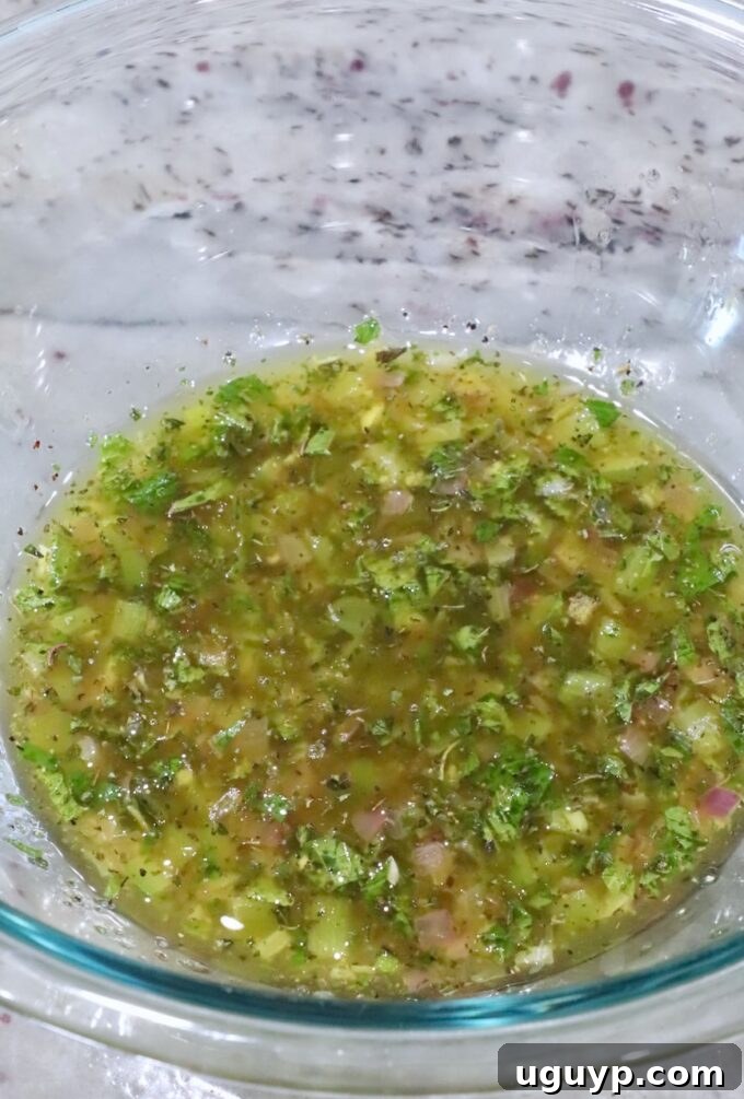A glass bowl containing whisked olive oil, lemon juice, red wine vinegar, mint, oregano, salt, and pepper, forming the base of the Greek dressing.