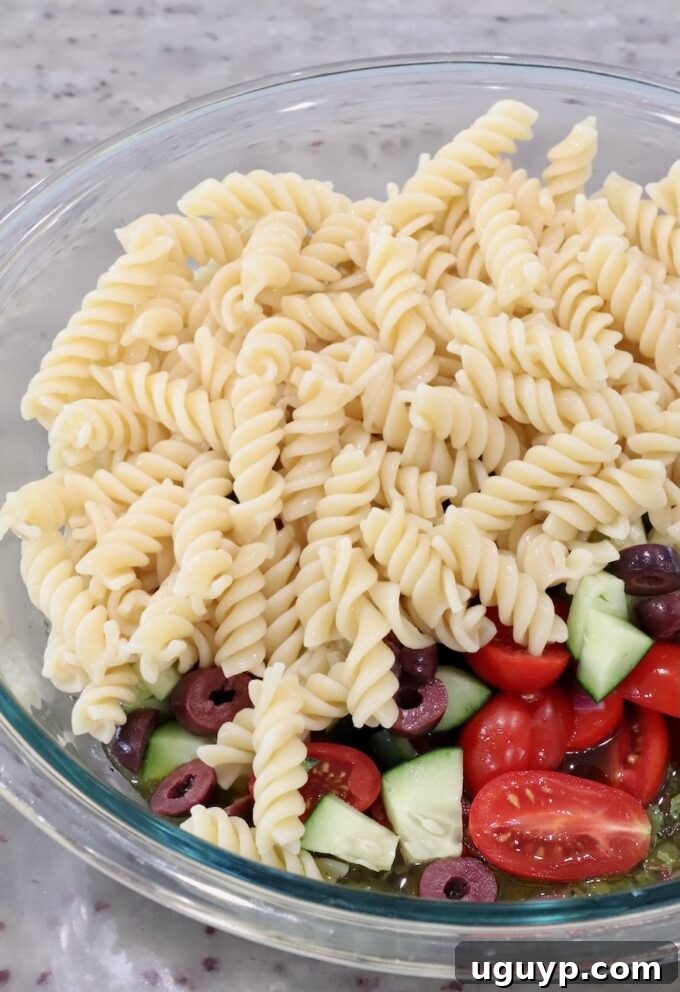 Cooked rotini pasta, diced cucumber, quartered cherry tomatoes, and sliced olives in a large glass bowl, ready to be combined with the dressing.