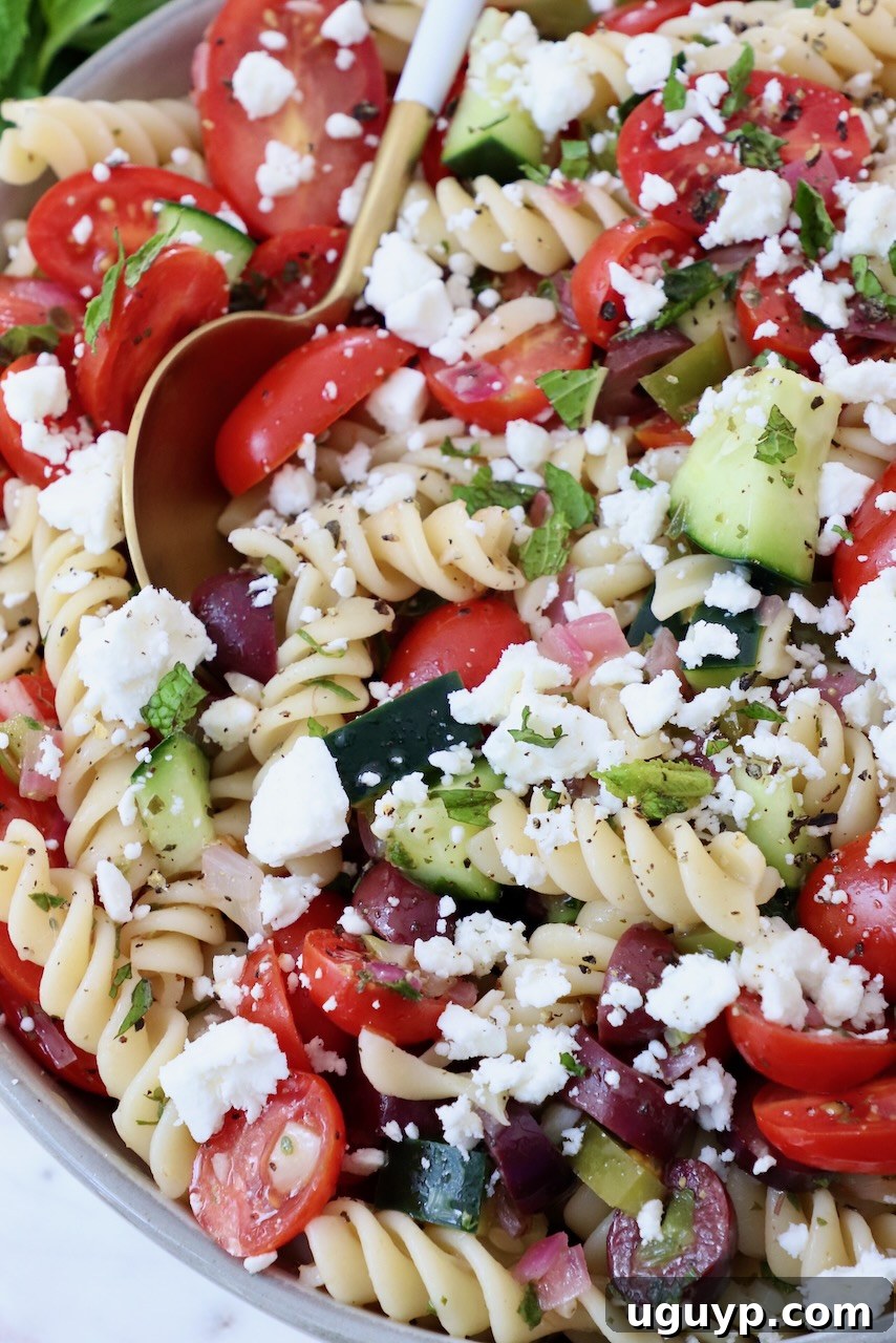 A close-up of Greek pasta salad topped with crumbled feta cheese in a white bowl, with a serving spoon.