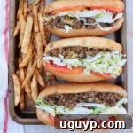 Three chopped cheese sandwiches, garnished with sliced tomatoes and shredded lettuce, artfully arranged on a baking sheet next to a pile of seasoned, golden French fries.