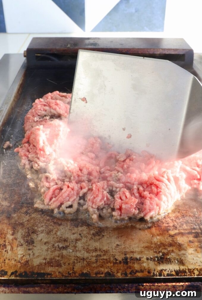 Close-up of ground beef sizzling on a hot flat top griddle, with a large metal spatula poised to chop and cook the meat.