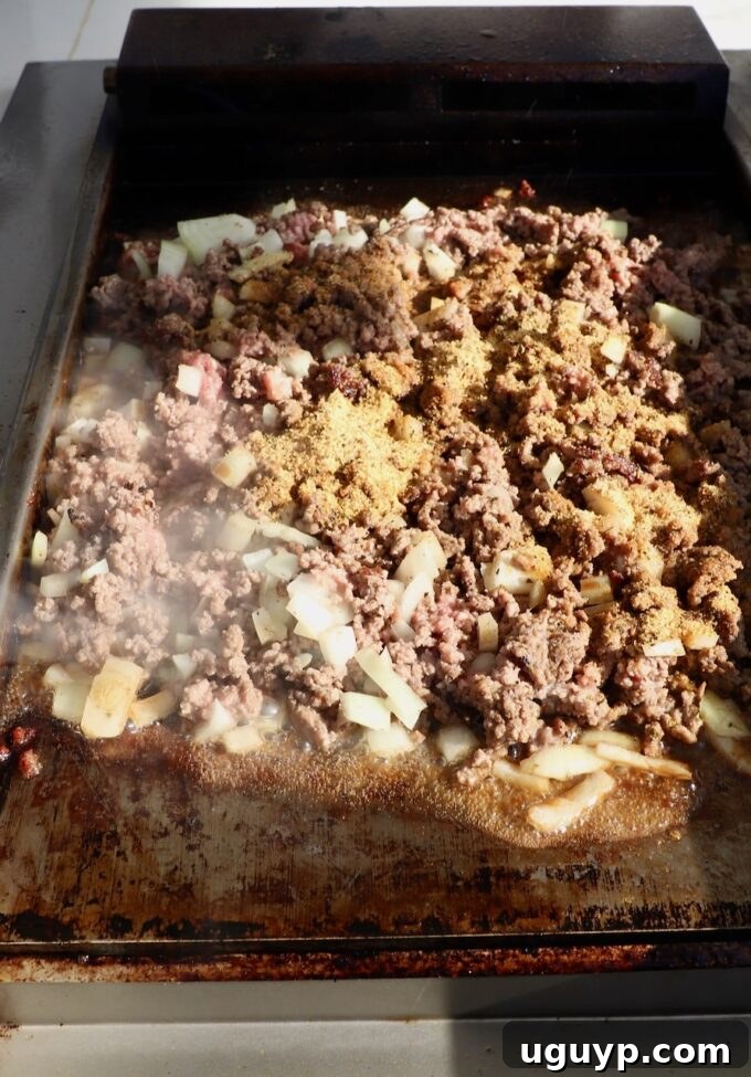 Perfectly seasoned ground beef and diced onions cooking and mingling on a flat top griddle, releasing aromatic steam and flavor.