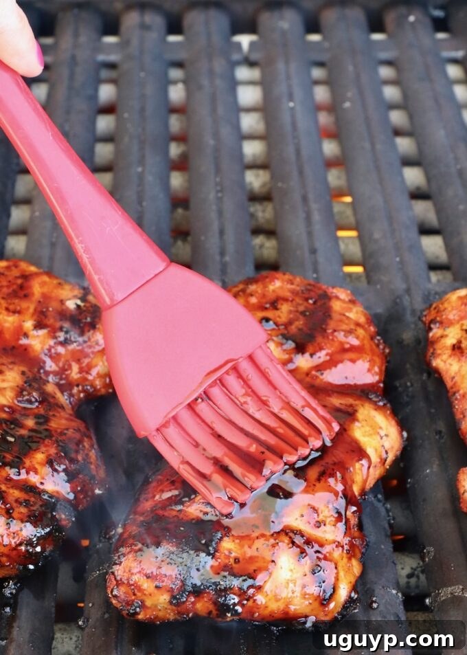 A basting brush applying a rich, red Korean BBQ sauce onto a sizzling grilled chicken thigh, creating a glossy glaze.