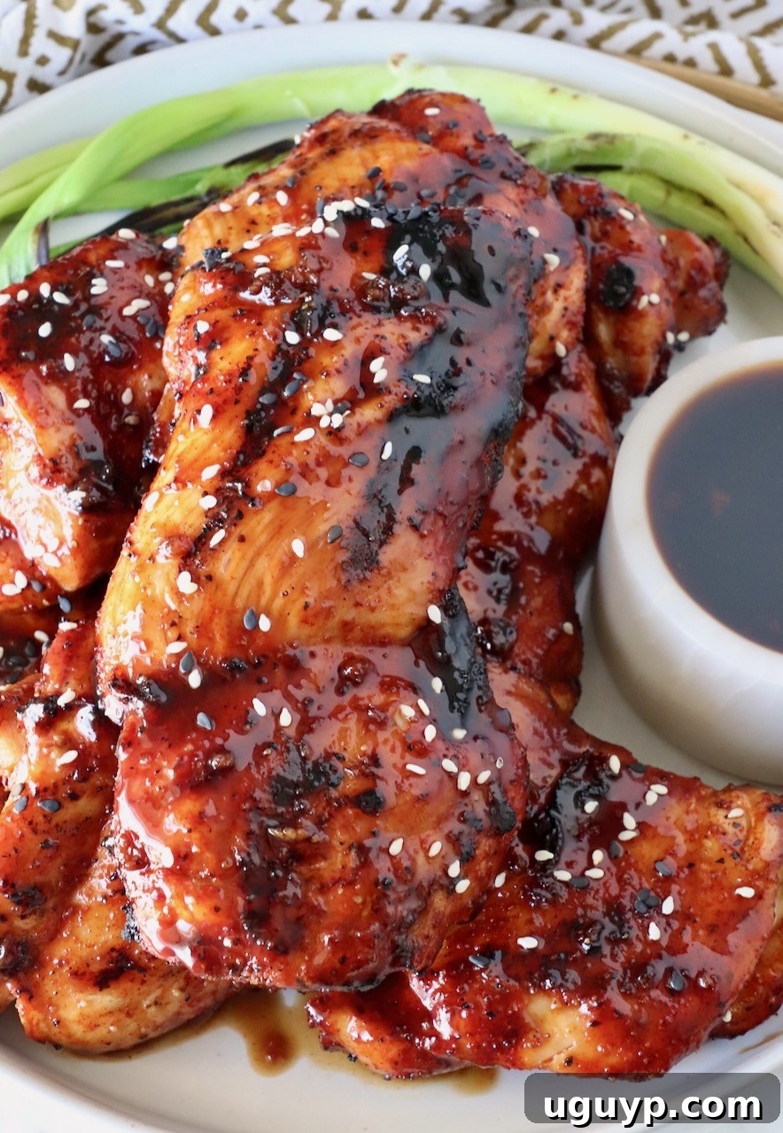Beautifully grilled Korean BBQ chicken thighs stacked on a rustic plate, glazed with rich sauce, accompanied by fresh green onions and a small bowl of extra Korean BBQ sauce.