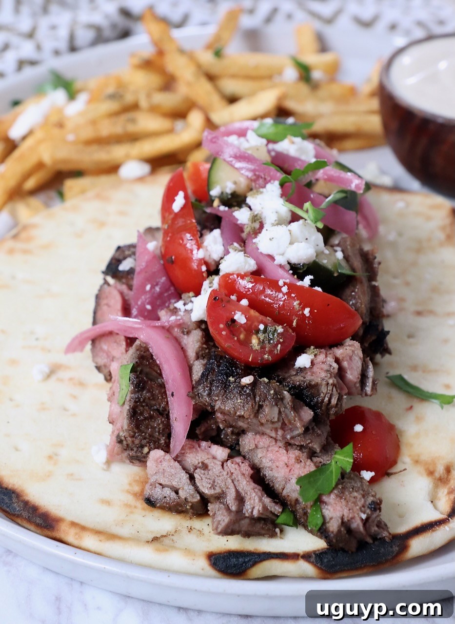 sliced, grilled beef shawarma on top of pita bread, garnished with tomatoes, red onions, and feta cheese, served on a plate with crispy french fries