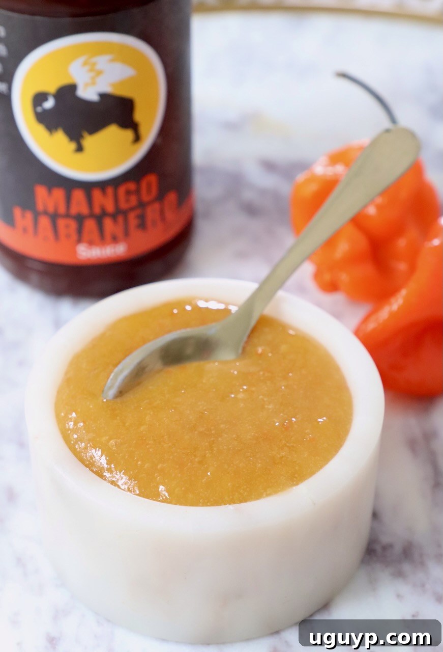 A vibrant bowl of homemade mango habanero sauce with a spoon, flanked by two fresh habanero peppers and a bottle of store-bought sauce in the background, symbolizing the inspiration for this copycat recipe.
