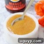 A small bowl of mango habanero sauce with a spoon, next to fresh habanero peppers and a bottle of store-bought sauce.