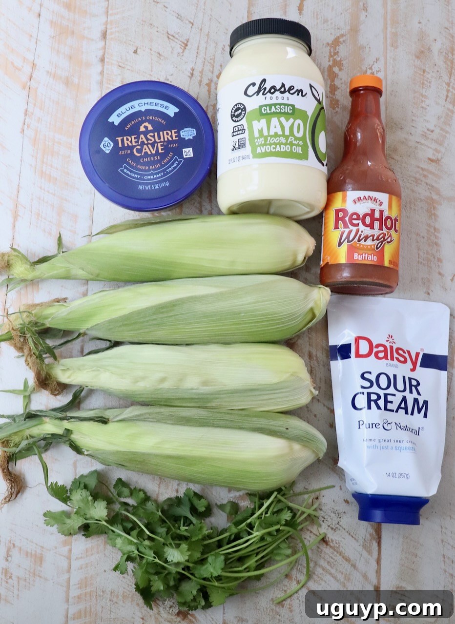 Various fresh ingredients for making Buffalo Elote laid out on a white wooden board, including corn on the cob, mayonnaise, sour cream, buffalo sauce, blue cheese crumbles, and fresh cilantro.