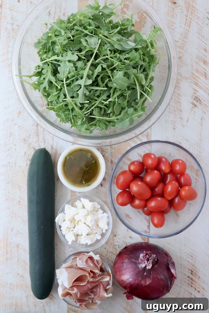 Assortment of fresh ingredients for cucumber prosciutto salad laid out on a white wooden board, including arugula, cucumber, tomatoes, red onion, prosciutto, and feta