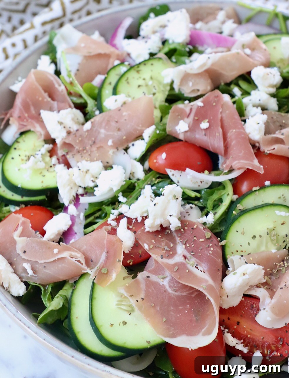 Close-up of prosciutto, cucumber slices, and tomatoes beautifully arranged on top of a bed of arugula in a bowl with feta crumbles