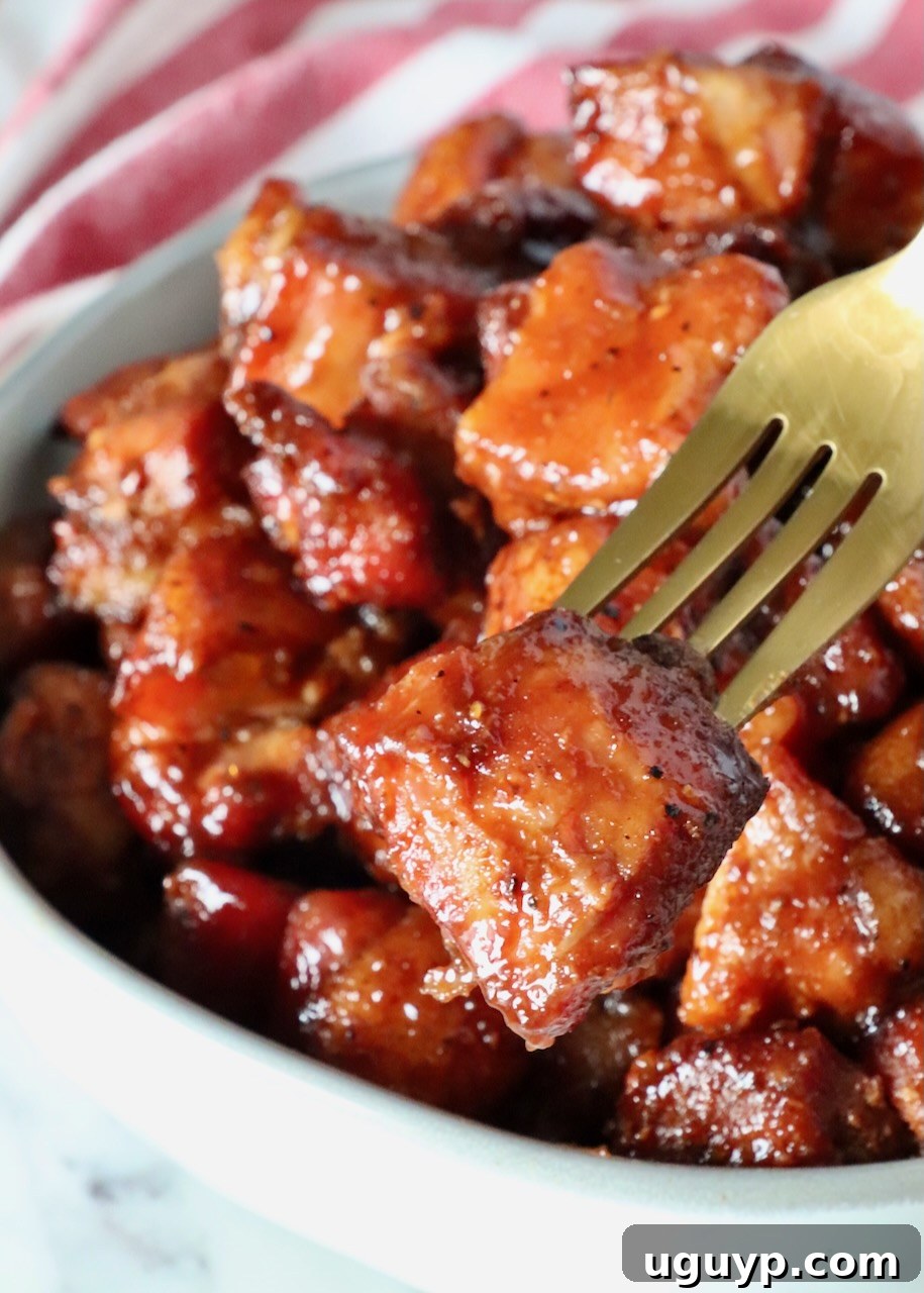 Fork in a pork belly burnt end held over a bowl of smoked pork belly cubes.