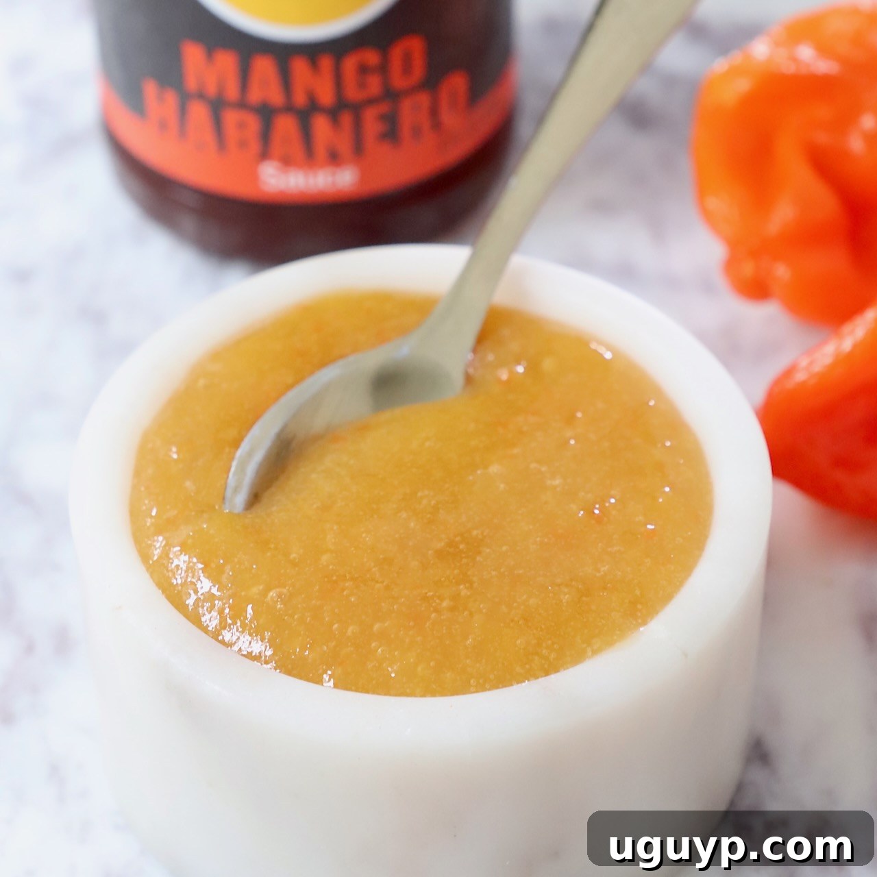mango habanero sauce in a bowl with a spoon with a bottle of sauce in the background and fresh habanero peppers