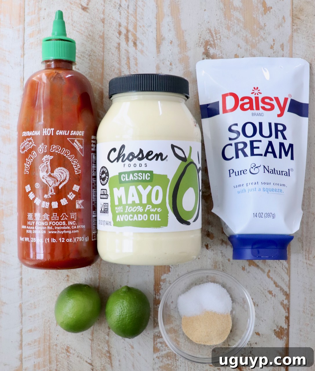 Condiments and ingredients on a wooden surface: a bottle of Sriracha hot sauce, avocado oil mayo, sour cream, two limes, and a small bowl of salt and garlic powder.