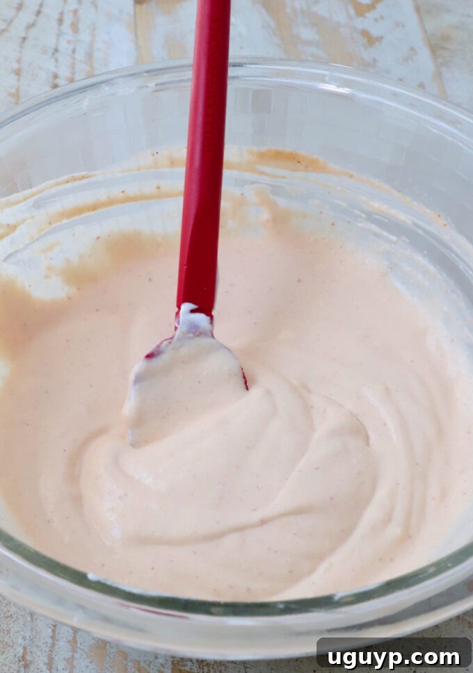 A clear glass bowl filled with creamy orange sauce, being mixed with a red spatula.