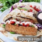 Three tacos with crispy fish, purple cabbage, diced tomatoes, and creamy sauce on a tortilla. With fresh cilantro in the background