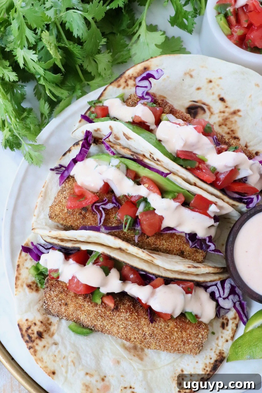 Three tacos with crispy fish, purple cabbage, diced tomatoes, and creamy sauce on a tortilla. They're garnished with fresh cilantro.