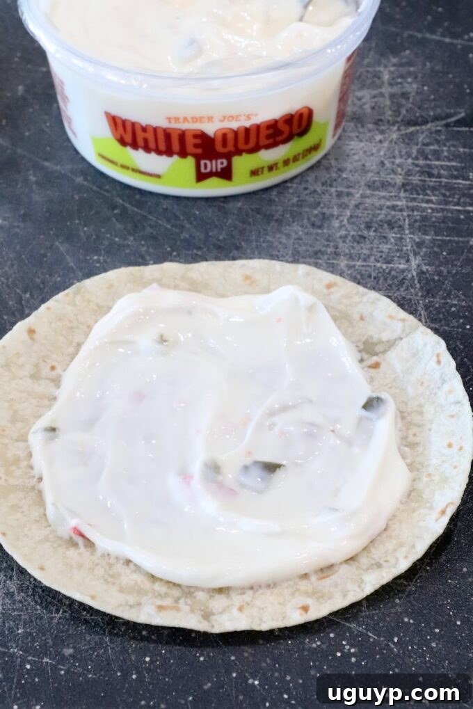 A small flour tortilla evenly spread with a creamy layer of white queso dip, preparing for the delicious filling.