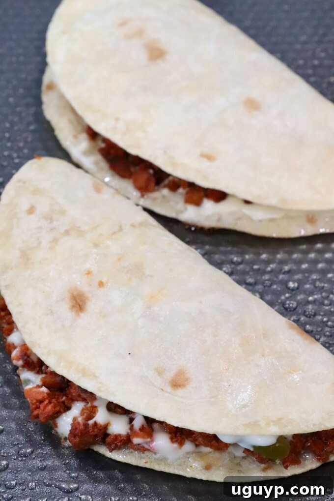 Two neatly folded soft tacos, generously filled with a mixture of cooked chorizo and melted cheese, awaiting their final crisping in the pan.