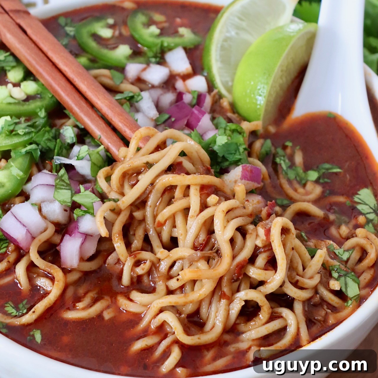 Cooked ramen noodles being lifted with chopsticks from a bowl of rich Birria broth, showcasing the delicious fusion dish.