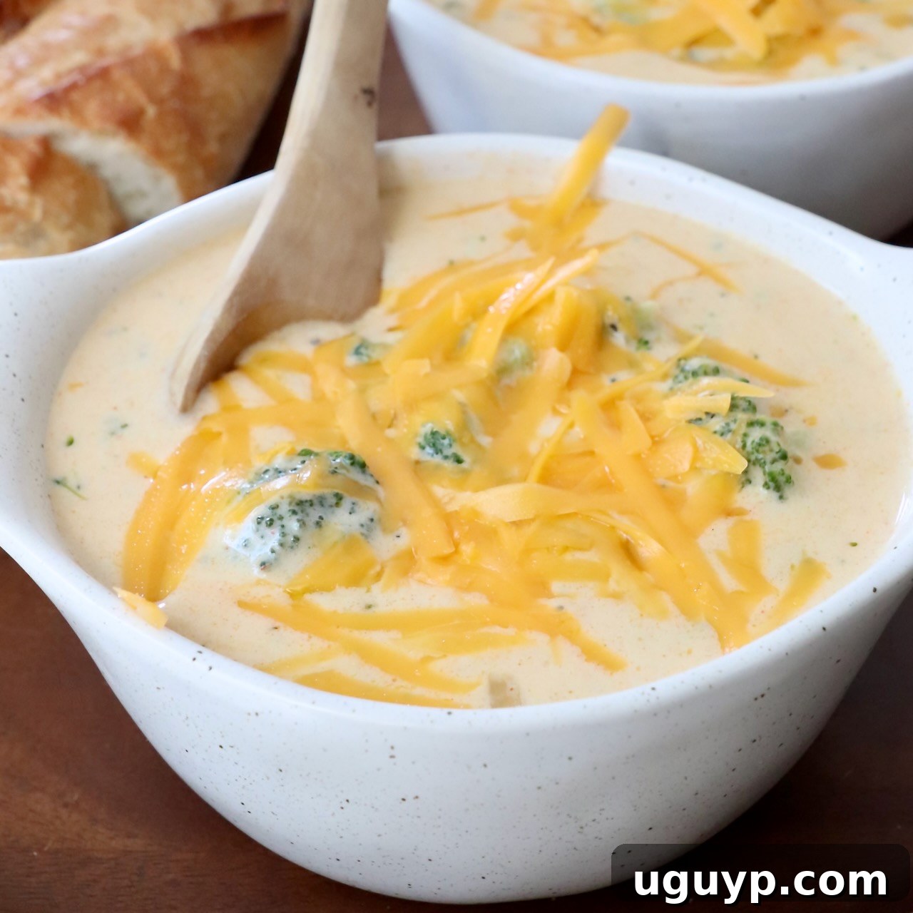 Rich Broccoli Cheddar Soup in a bowl, generously topped with shredded cheddar cheese and a wooden spoon resting in it.