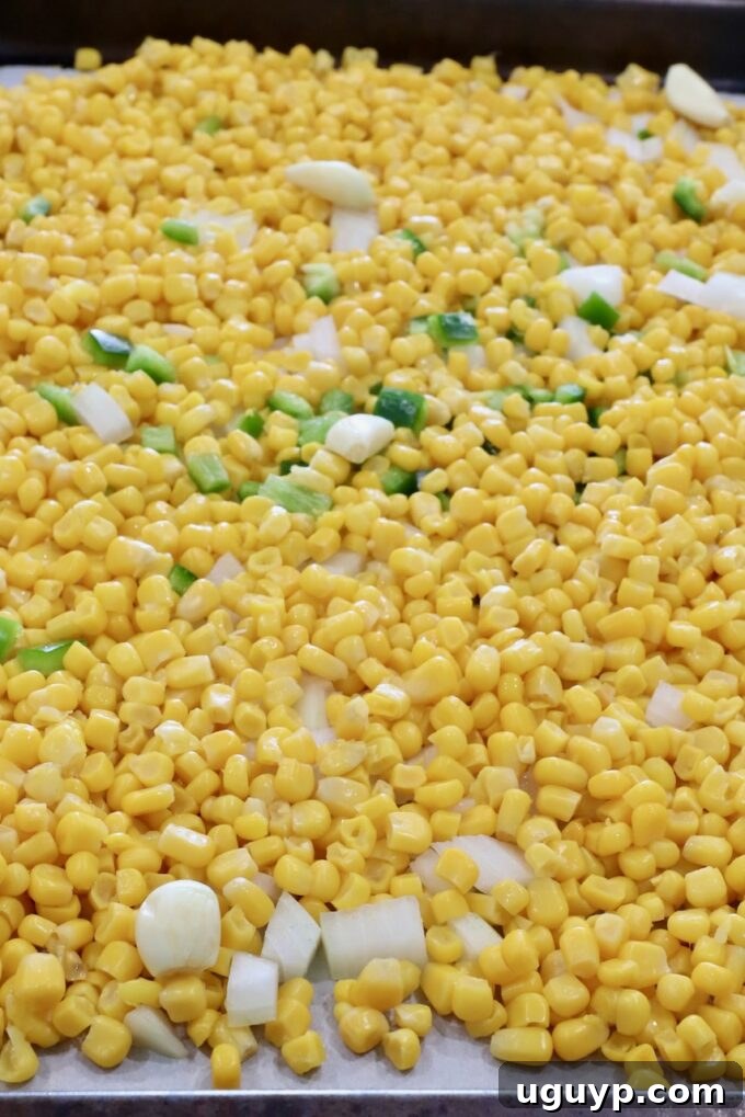 Corn kernels mixed with diced onions, jalapeños, and whole garlic cloves, spread evenly on a baking sheet, ready for seasoning and roasting.
