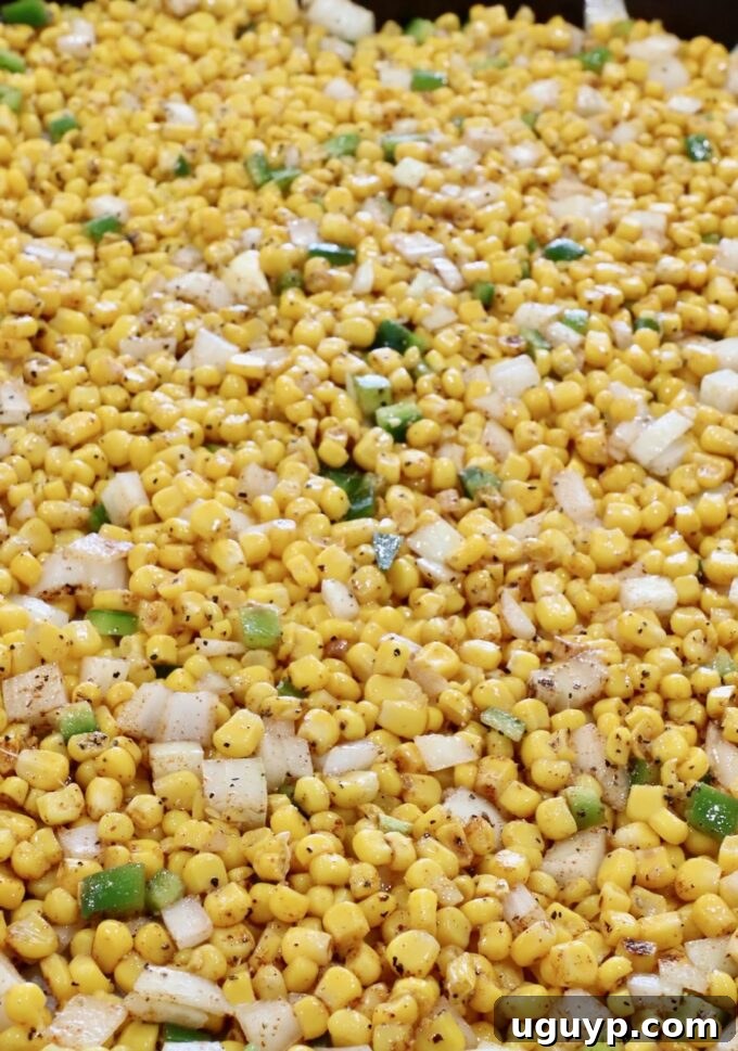 A close-up view of seasoned corn kernels, diced onions, jalapeños, and whole garlic cloves on a baking sheet, coated in olive oil and spices, ready for the oven to begin roasting.