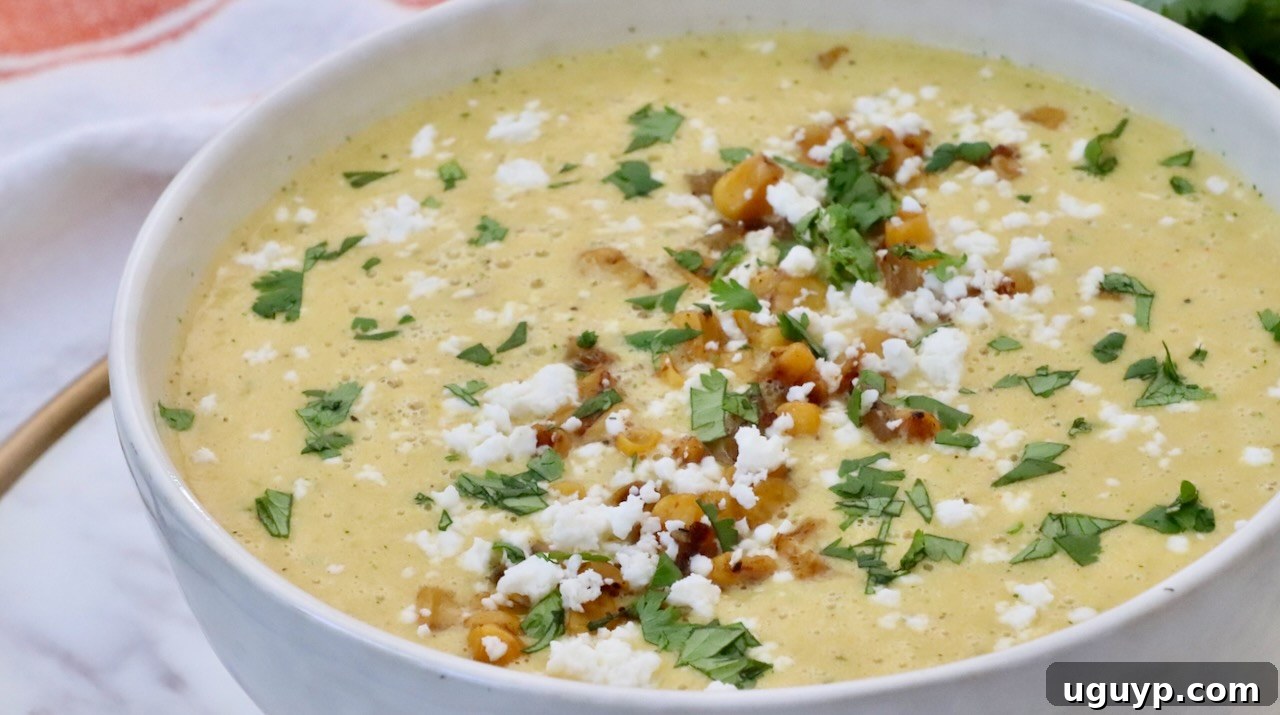 A close-up of a white bowl overflowing with creamy Mexican Street Corn Soup, beautifully garnished with bright green cilantro, more crumbled cotija cheese, and golden roasted corn kernels, highlighting its inviting texture and rich appeal.