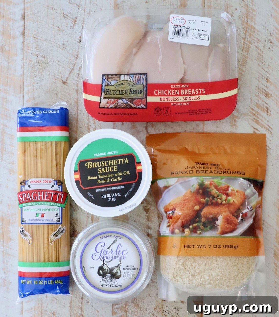 A selection of Trader Joe's products on a light wooden surface, including chicken breasts, bruschetta sauce, spaghetti, garlic spread, and panko breadcrumbs.