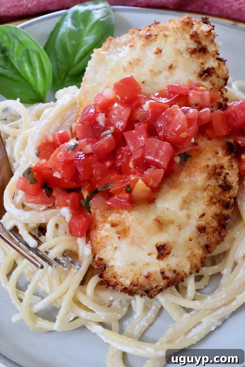 piece of crispy chicken on top of pasta on a plate, topped with tomato bruschetta, with basil leaves in the background