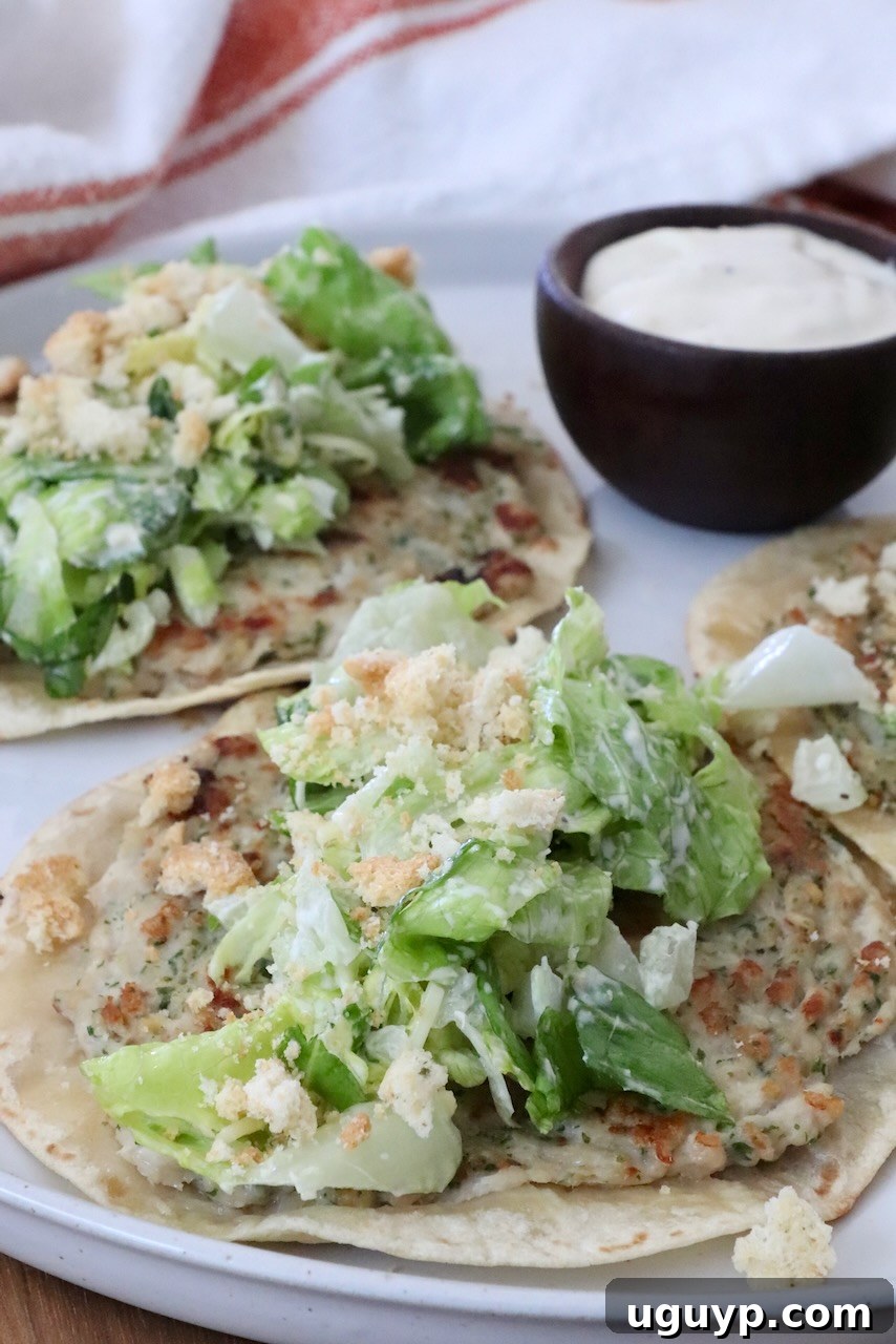 Two crispy chicken caesar smash tacos, garnished with fresh leafy green salad and crunchy croutons, artfully arranged on a pristine white plate. A small, elegant bowl of creamy Caesar dressing sits beside them, ready for dipping.