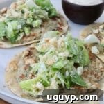 Two smash chicken tacos topped with leafy green salad and crumbled croutons on a white plate. A small bowl of creamy dipping sauce is on the side.