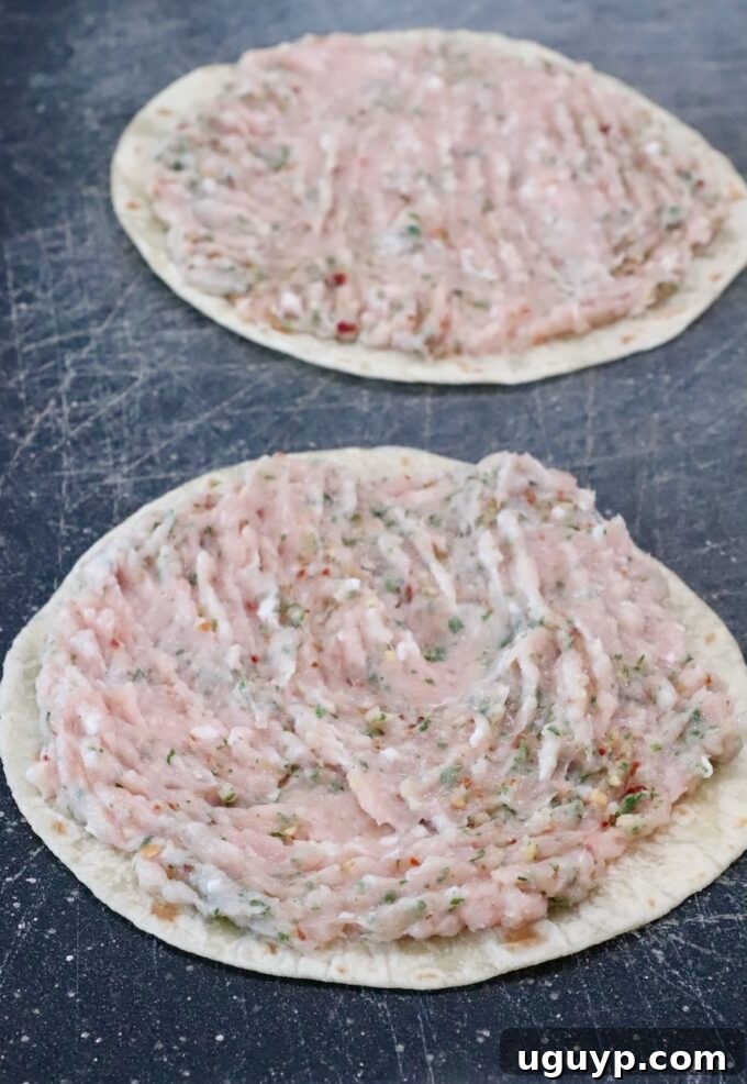 Two soft tortillas laid flat on a dark cutting board, each generously topped with a mound of raw ground chicken that has been seasoned with herbs and spices, ready for smashing and cooking.