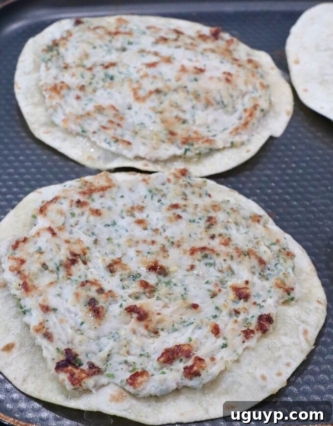 A close-up shot of two crispy chicken smash tacos cooking on a textured, dark griddle pan. The chicken, seasoned with herbs, is perfectly browned and adhered to the golden-brown tortillas.