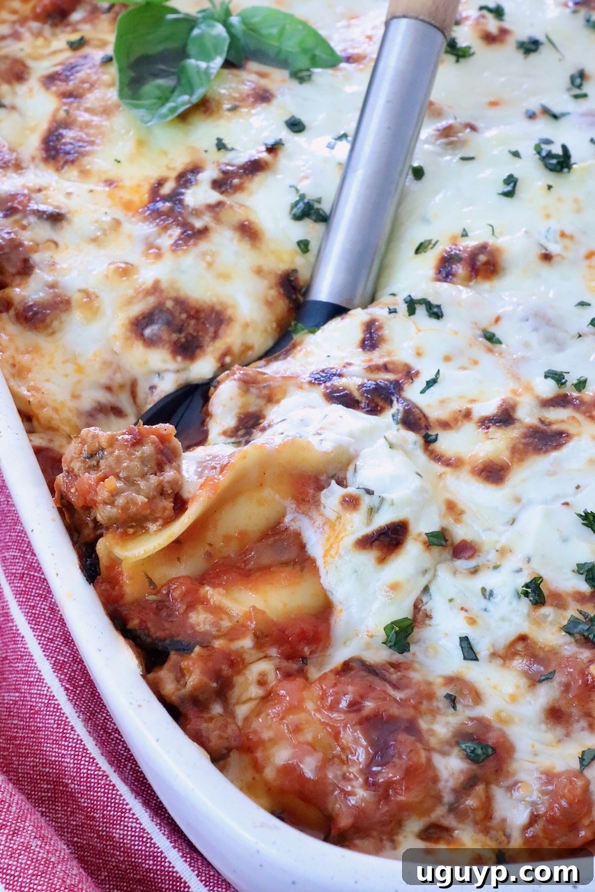 A close-up view of a baked ravioli casserole, golden brown and bubbling, in a white ceramic dish. A serving spoon lifts a generous portion, revealing layers of pasta, rich meat sauce, and creamy cheese, garnished with fresh green herbs.
