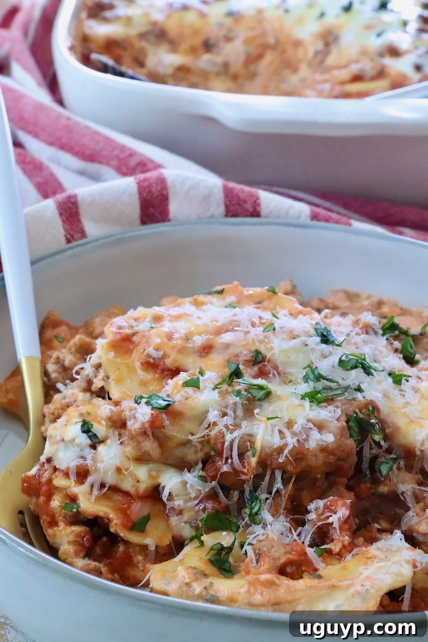 A close-up shot of a single serving of baked ravioli on a white plate, topped with generous melted cheese and garnished with fresh basil leaves. In the blurred background, a red-striped towel and the full casserole dish are visible.