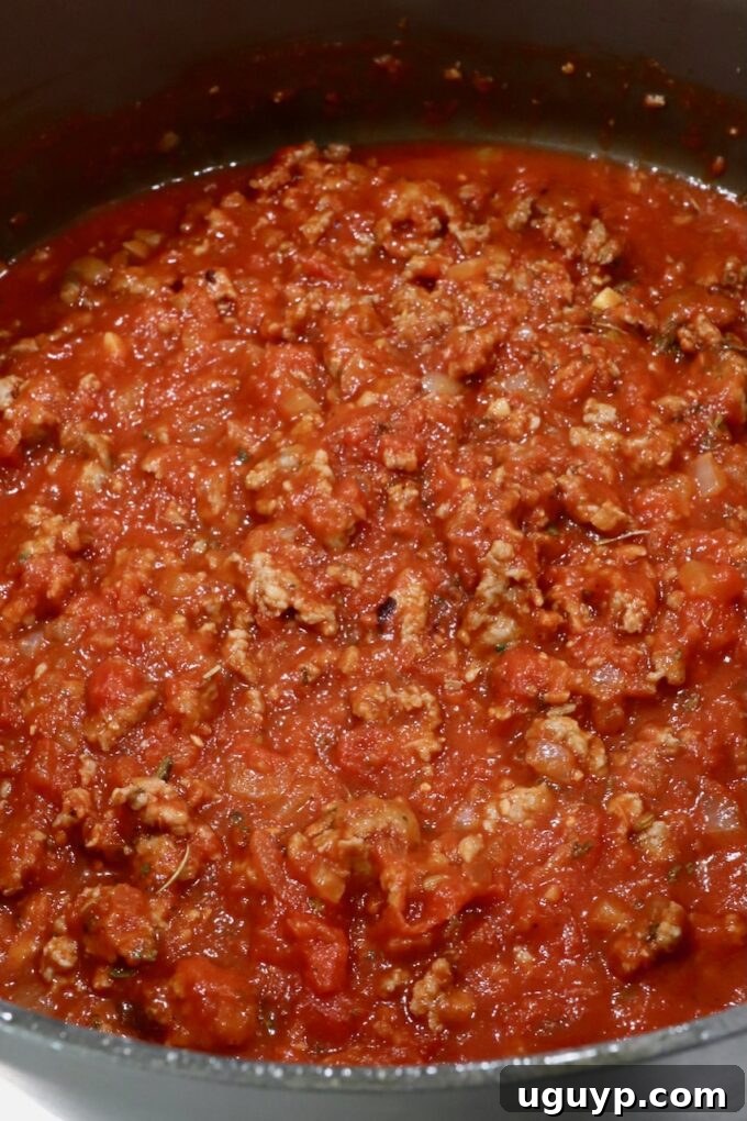 A vibrant red tomato meat sauce, rich with browned Italian sausage and aromatic herbs, simmers gently in a pot, ready to infuse its flavors into the ravioli casserole.
