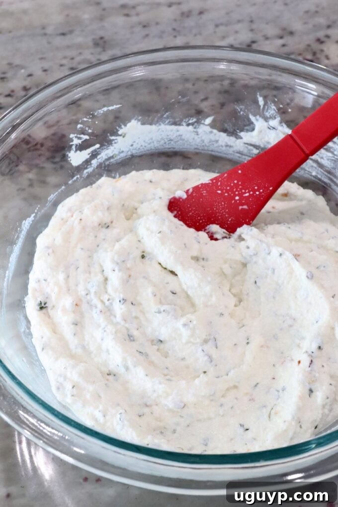 A bowl of creamy, white ricotta cheese, lightly seasoned with Italian herbs and a touch of mozzarella, sits on a marble countertop, prepared for layering in the baked ravioli.