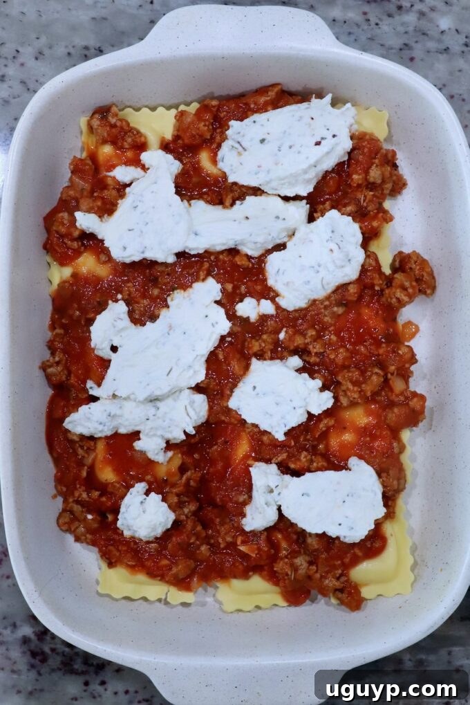 The first layer of ravioli in a baking dish is topped with a rich meat sauce and dollops of seasoned ricotta cheese, ready for the next layer of the casserole.