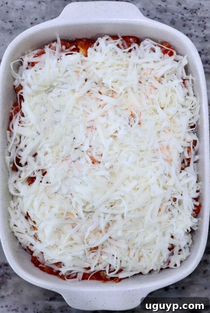 A casserole dish filled with layers of baked ravioli, meat sauce, and ricotta, generously topped with a final layer of shredded mozzarella cheese, ready for baking.