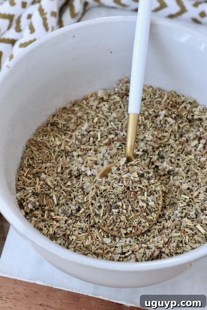 A white bowl filled with mixed dried herbs and spices, with a gold and white spoon resting inside. A patterned cloth is partially visible in the background.