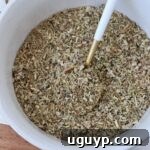 A bowl filled with a coarse, mixed herb blend featuring green and brown hues. A golden spoon with a white handle rests inside, evoking a rustic culinary vibe.