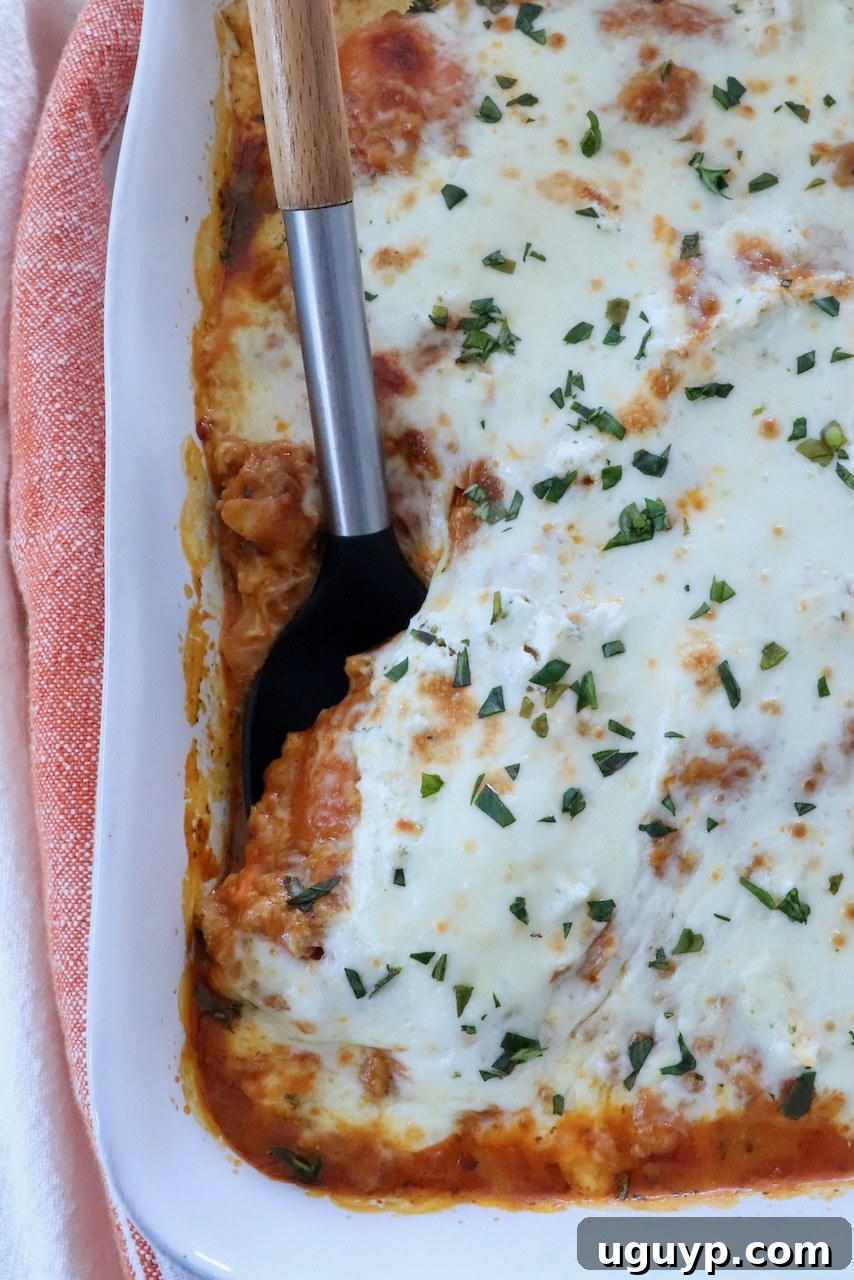 Close-up of golden-brown butternut squash baked ravioli in a white baking dish, featuring melted cheese sprinkled with fresh herbs. A black spatula has removed a serving, revealing the creamy layers beneath.