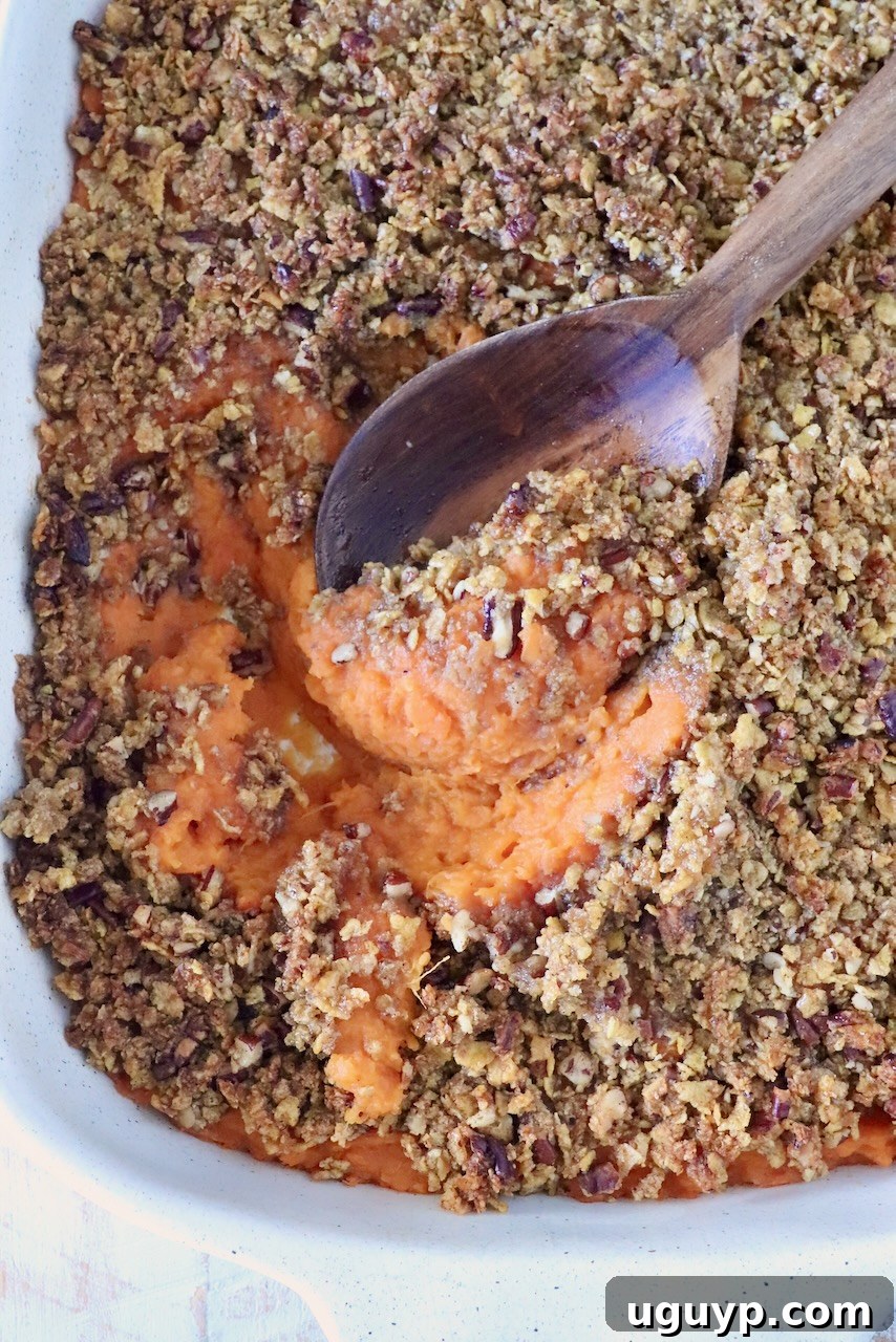 Close-up of a sweet potato casserole with a crunchy pecan topping and a wooden spoon scooping the orange sweet potato filling out from the dish