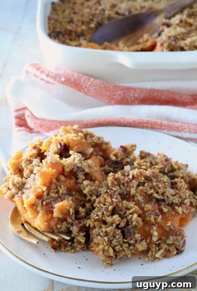 Plate of sweet potato casserole topped with a crispy pecan-crumb layer. A serving spoon lies nearby; a baking dish and towel are in the background.