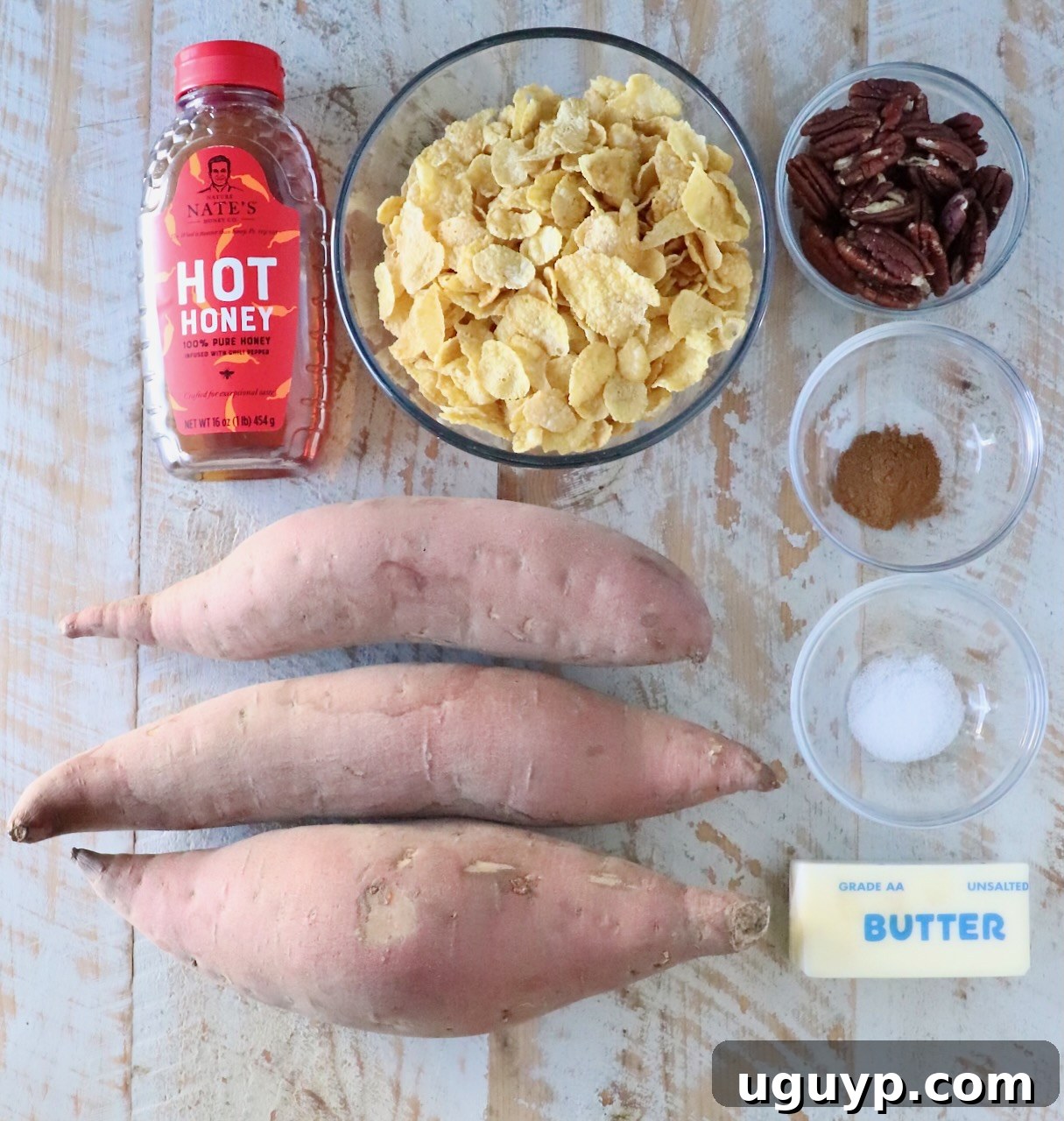 Ingredients for a sweet potato dish on a wooden surface: three raw sweet potatoes, a bottle of hot honey, cornflakes in a bowl, pecans, cinnamon, salt, and a stick of butter.