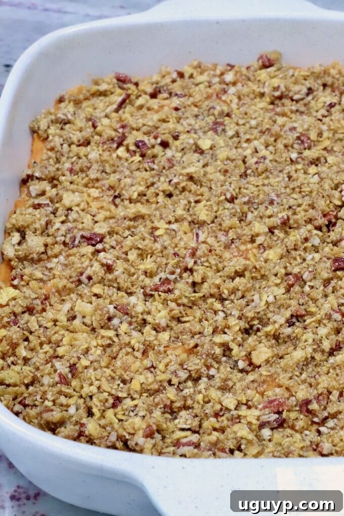 A white ceramic dish filled with a golden-brown, crumbly topping covering a sweet potato casserole, with visible pecan pieces throughout