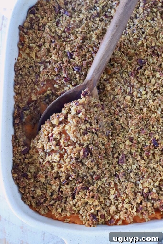 A white baking dish holds a sweet potato casserole topped with a crumbly pecan and corn flake crust. A wooden spoon scoops a portion, revealing orange filling.