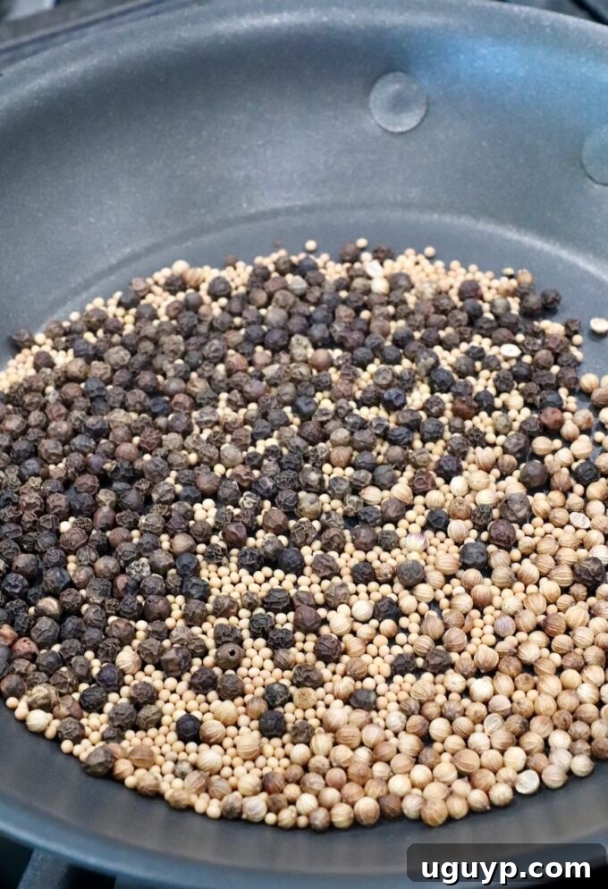 An array of distinct whole spices, including glossy black peppercorns, pale mustard seeds, and round coriander seeds, undergoing a delicate dry roasting process in a dark, cast-iron frying pan, releasing their rich aromas.