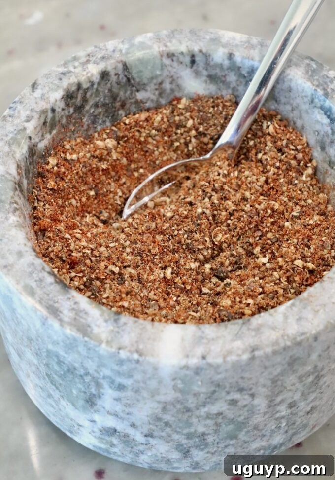 A detailed close-up of a sturdy marble mortar, filled with a freshly prepared, coarsely ground mixture of mixed spices. A gleaming metal spoon rests within the blend, highlighting the rustic texture of the seasonings.